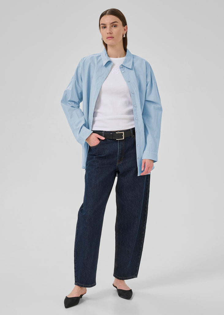 Woman wearing a light blue shirt, white top, dark jeans, and black shoes on a light gray background