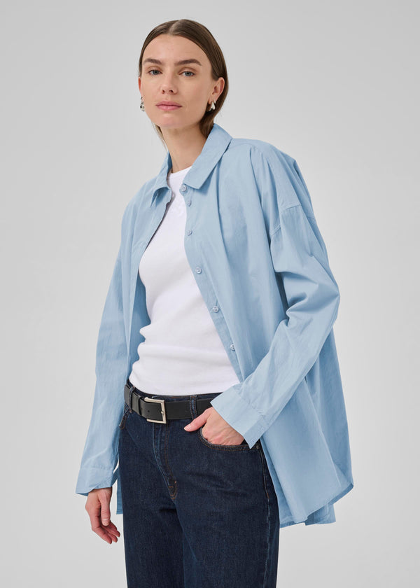 Woman wearing a light blue shirt over a white top with dark jeans on a plain background