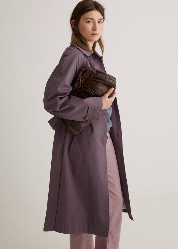 Woman wearing a long purple coat holding a brown leather bag against a plain background