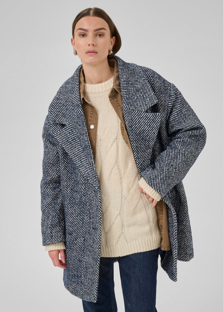 Woman wearing a blue herringbone coat over a cream sweater and brown shirt on a gray background