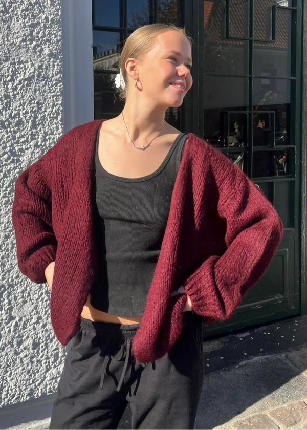Woman wearing a red cardigan over a black top, standing against a building with large windows.