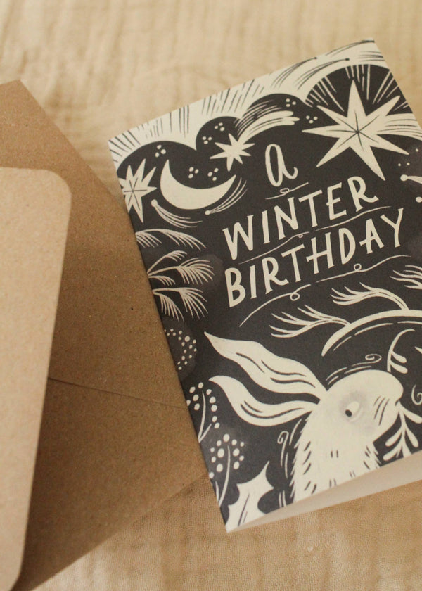 Greeting card with 'A Winter Birthday' text and winter-themed illustrations on a wooden surface.