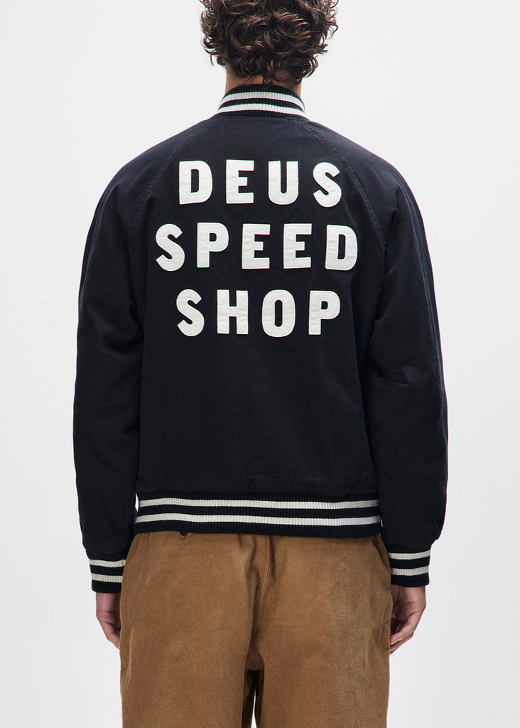 Person wearing a black jacket with 'DEUS SPEED SHOP' text on the back against a white background