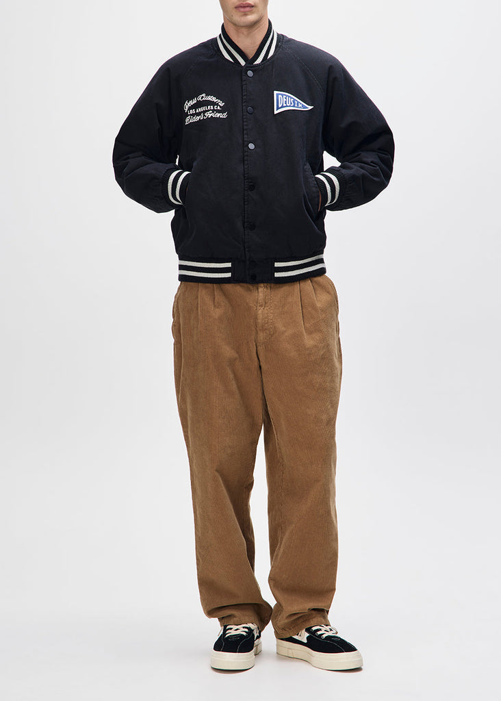 Person wearing a black varsity jacket with white accents and brown pants on a white background