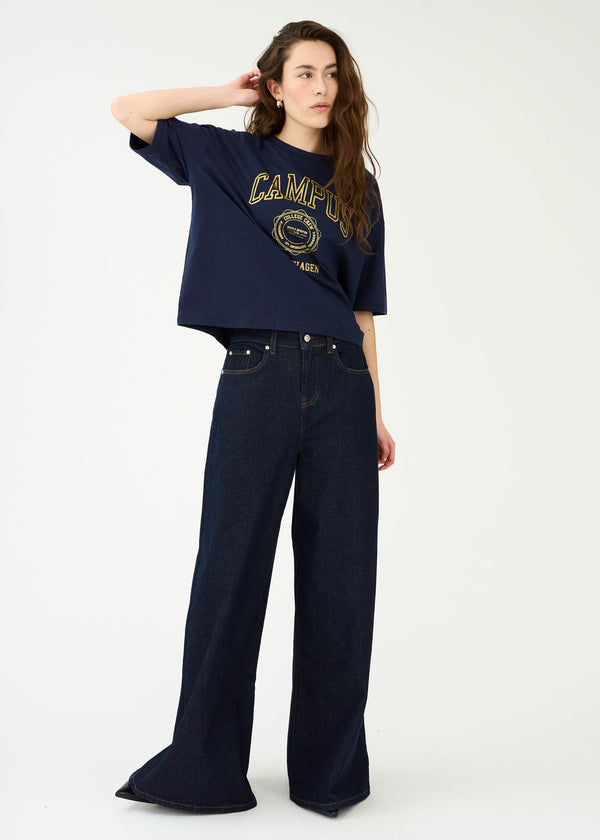 Woman wearing a navy blue oversized t-shirt with 'Camper' logo and dark jeans on a white background