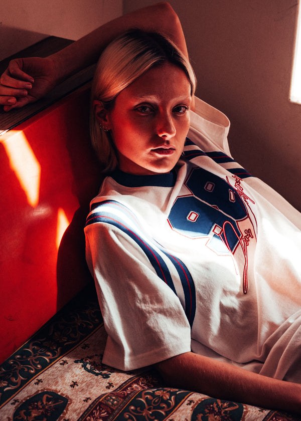 Person wearing a white shirt with a blue emblem, leaning against a red wall.