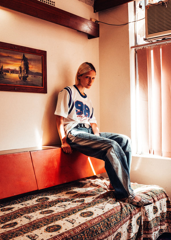 Person sitting on a patterned bed in a room with a painting on the wall and a window.