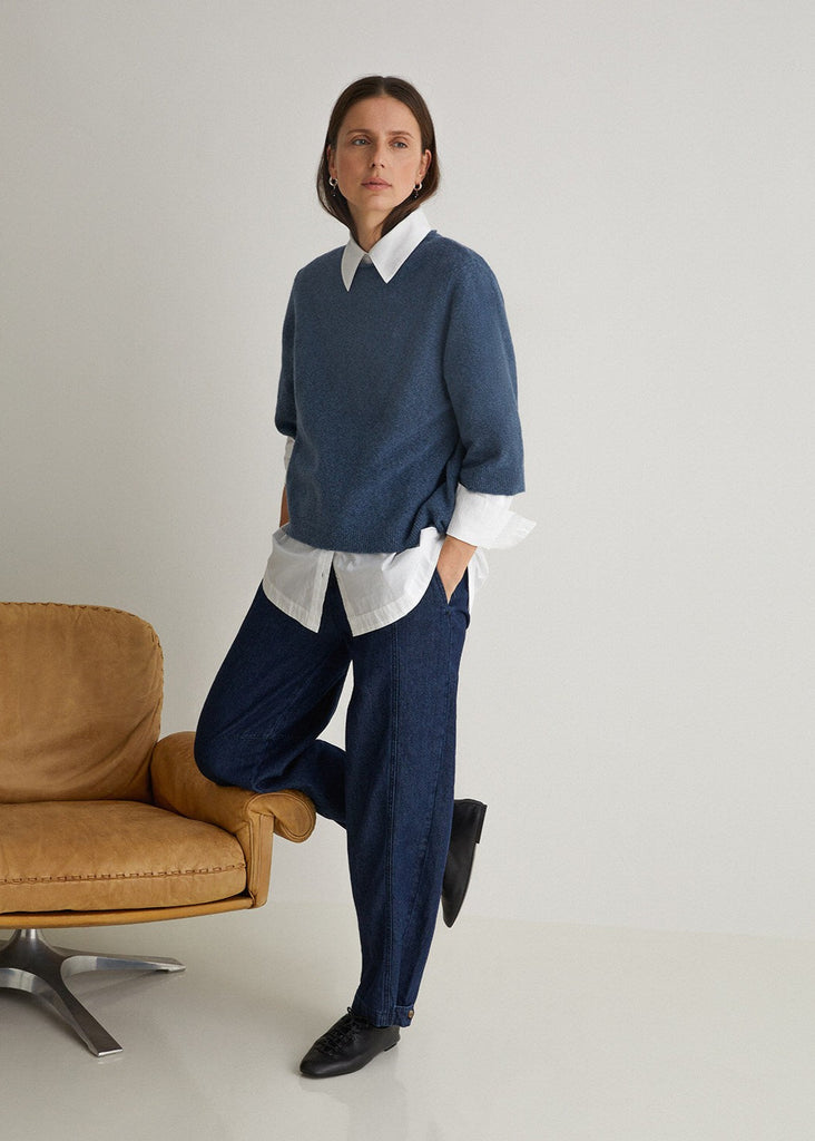 Woman wearing a blue sweater over a white shirt with blue pants, standing in a minimalistic room.