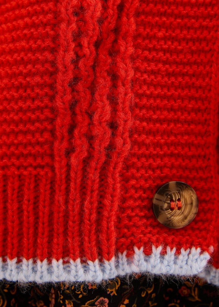 Close-up of a bright red knitted garment, showing a ribbed cuff with a white stripe and a small round brown button, with a floral patterned fabric visible along the bottom edge.