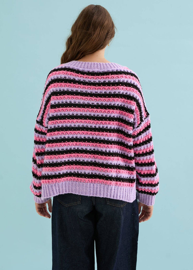 A woman viewed from behind wearing a chunky knit sweater with horizontal stripes in lavender, pink, and black, paired with dark blue jeans against a solid light blue background.