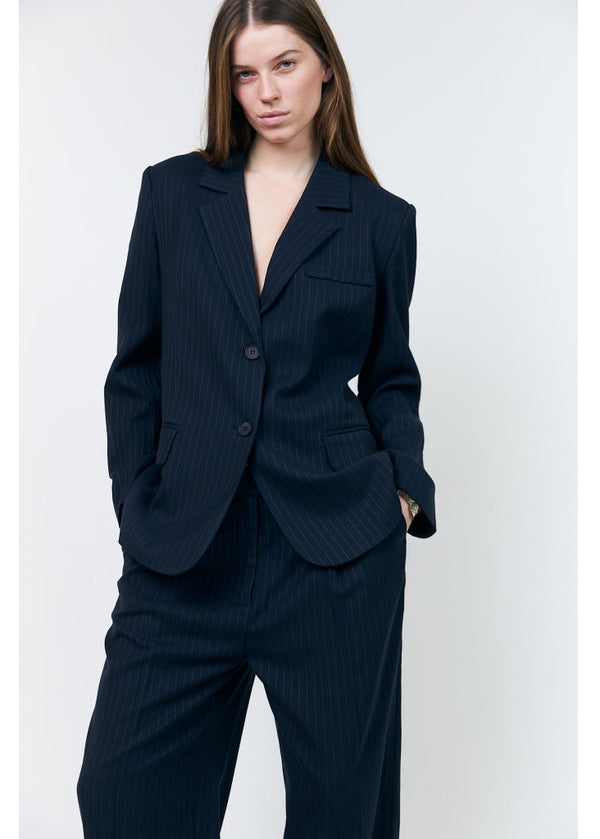 A woman modeling a dark navy pinstripe blazer and matching trousers against a light gray studio background.