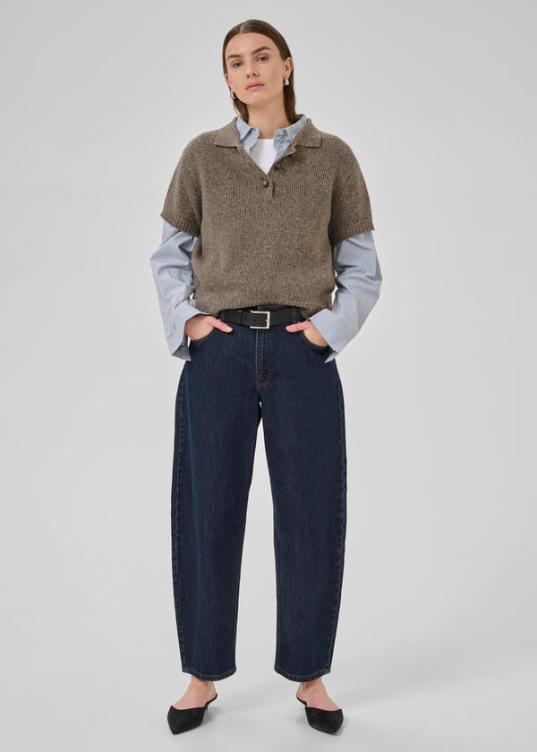 Woman wearing a brown sweater over a light blue shirt with dark jeans on a white background
