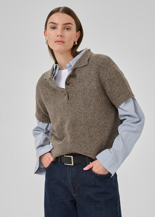 Woman wearing a brown knitted sweater over a light blue shirt with dark jeans on a plain background
