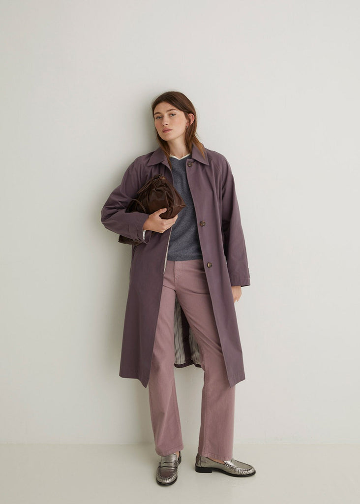 Woman wearing a long purple coat holding a brown bag against a white background