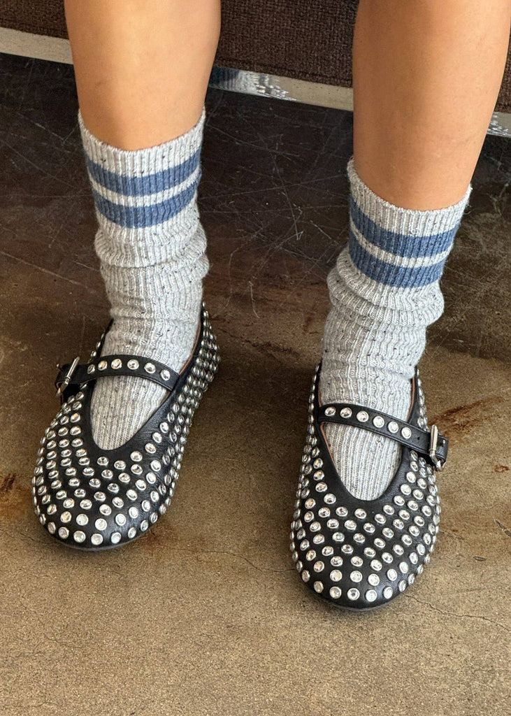 Black studded shoes worn with gray socks featuring blue stripes on a textured floor.