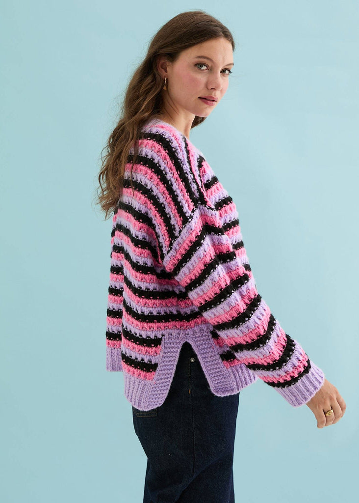 A woman wearing a pink, black, and light purple striped crochet cardigan, photographed from the side against a solid light blue background.