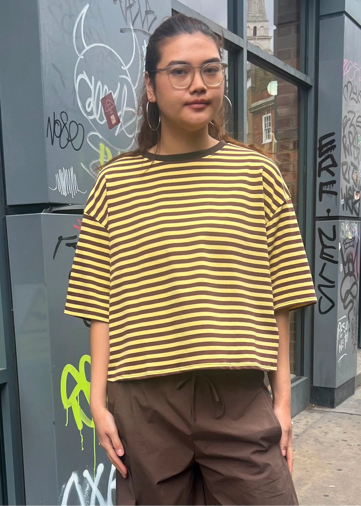 Person wearing a yellow and black striped shirt and brown pants standing in front of a graffitied wall.