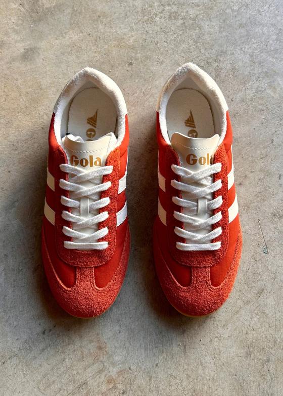 A pair of red suede Gazelle sneakers with white leather side stripes, white laces, and cream-colored interiors, displayed on a textured concrete floor.