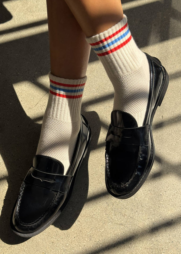 Black loafers worn with white socks featuring red and blue stripes on a concrete surface.