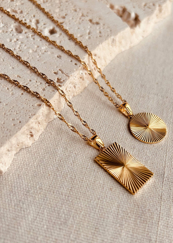 Two gold necklaces with geometric pendants on a textured beige surface