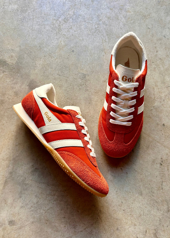 A pair of retro-style sneakers in red and white, photographed on a textured gray concrete floor. The shoes feature red suede and mesh panels, white side stripes, white laces, and a beige gum sole.