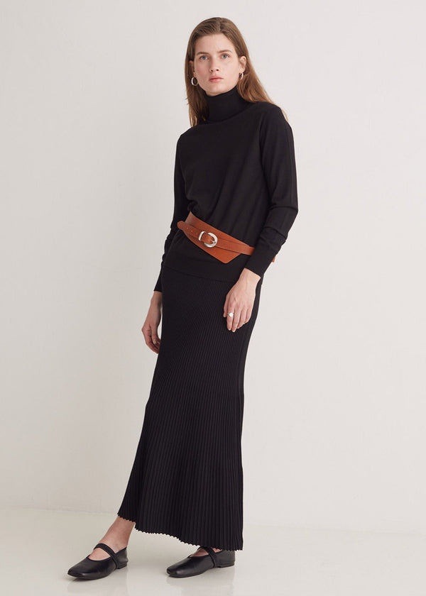 Woman wearing a black turtleneck and long skirt with a brown belt on a white background