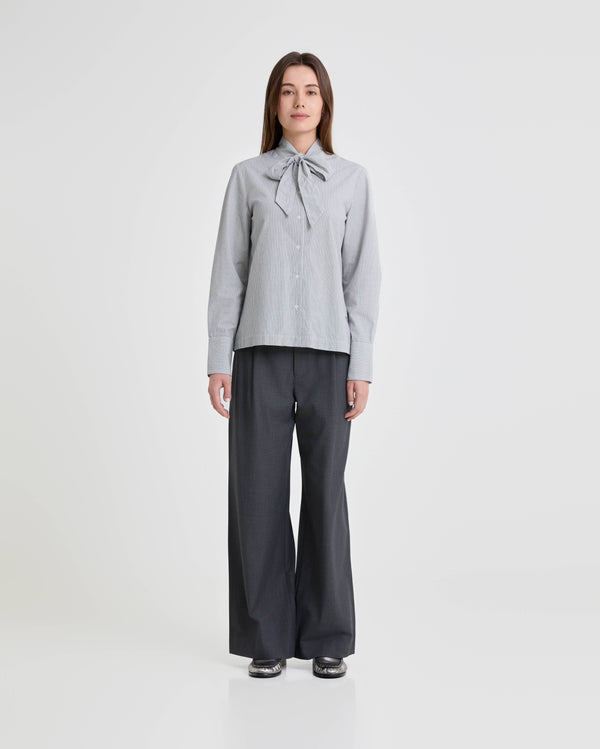 Person wearing a light gray checkered blouse with a bow and dark gray wide-leg pants on a white background