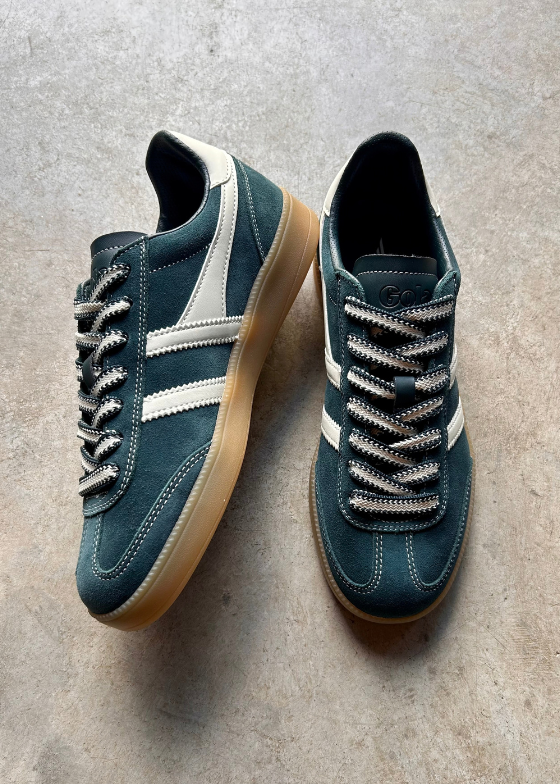 A pair of low-top sneakers in dark teal suede with off-white side stripes, cream laces, and gum soles, displayed on a textured gray concrete floor.