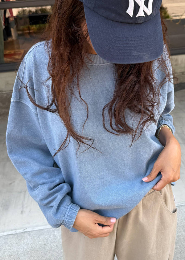 Person wearing a blue sweatshirt and cap with a logo, standing on a street.