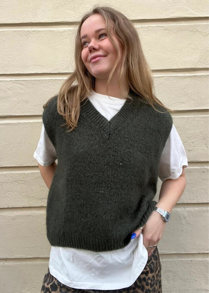 Person wearing a dark green knitted vest over a white shirt with a beige wall background