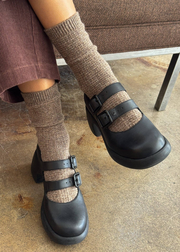 Black clogs with brown socks worn on a concrete floor.