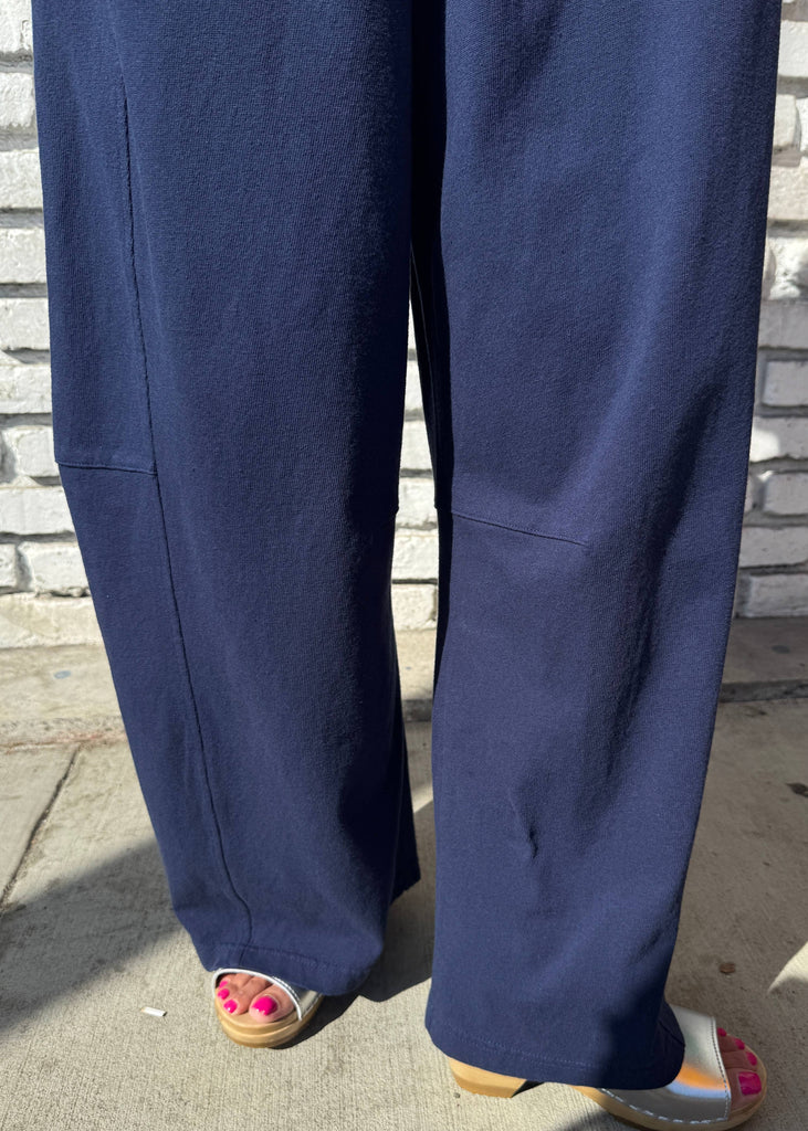 Close-up of a person wearing wide-leg navy blue pants and colorful slide sandals, standing outdoors against a white brick wall.