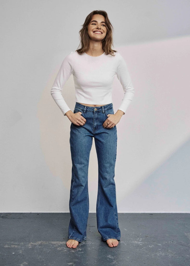 Woman wearing a white long-sleeve top and blue jeans standing against a plain background