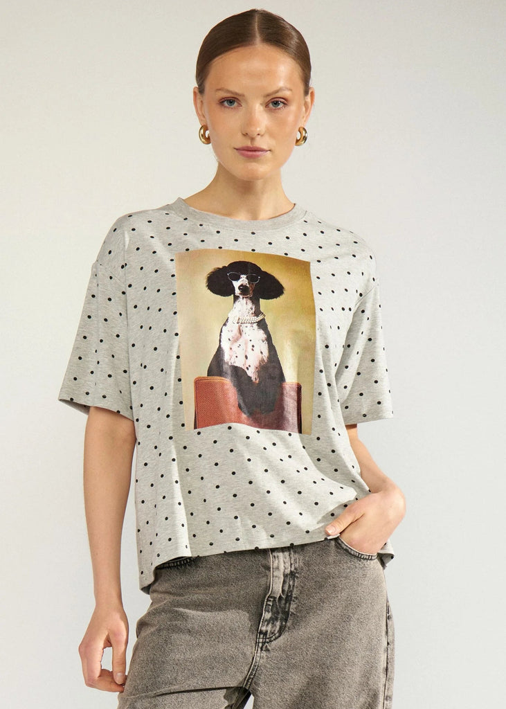 A woman modeling a light gray oversized t-shirt with black polka dots and a large graphic print of a black and white dog on the front, paired with gray jeans against a light gray studio background.