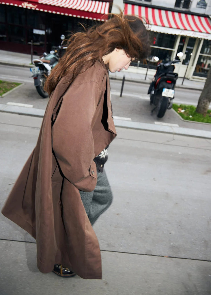 Person wearing a long brown coat on a city street with motorcycles and buildings in the background.
