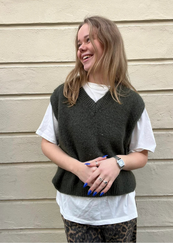 Woman wearing a dark green vest over a white shirt against a beige wall.