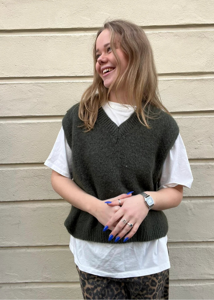 Woman wearing a dark green vest over a white shirt against a beige wall.