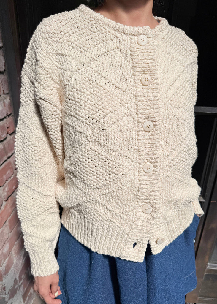 Person wearing a cream knitted sweater and blue pants against a brick wall.