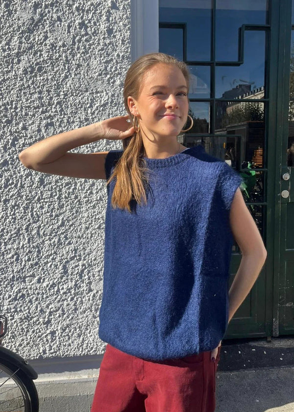 Woman wearing a blue sleeveless top and red pants standing against a textured wall.