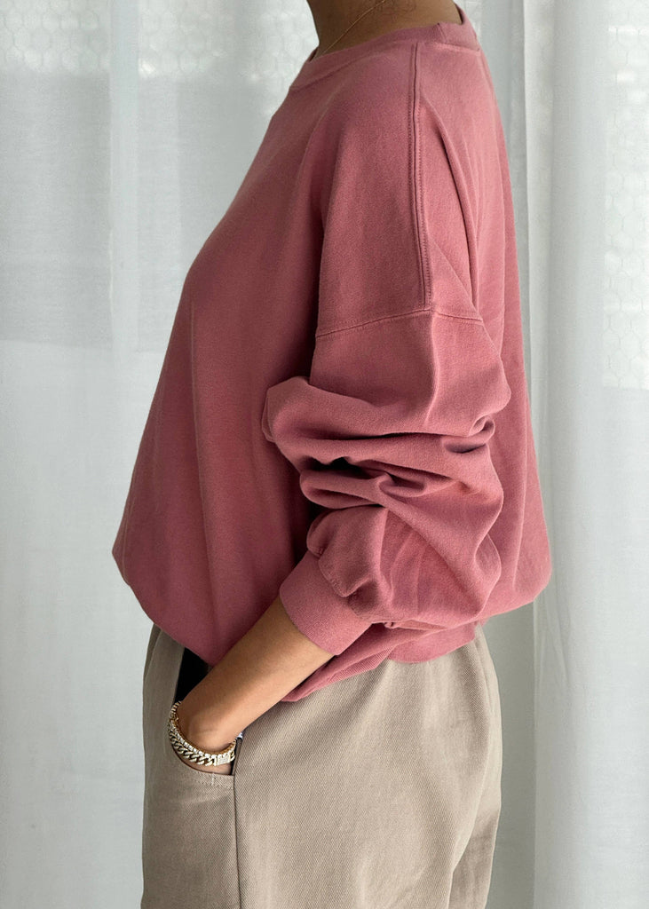 Person wearing a pink sweatshirt and beige pants against a white curtain background