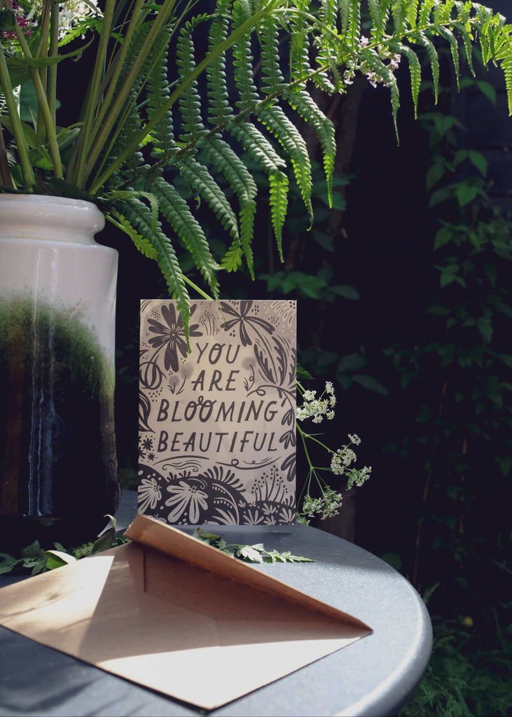 Decorative sign with 'You are blooming beautiful' text on a table with plants in the background