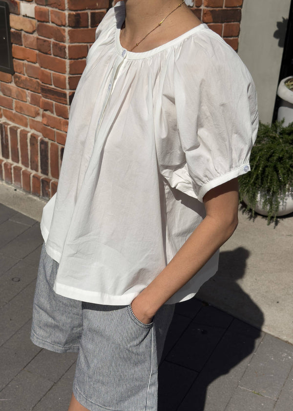 Person wearing a white blouse and gray shorts standing on a sidewalk.
