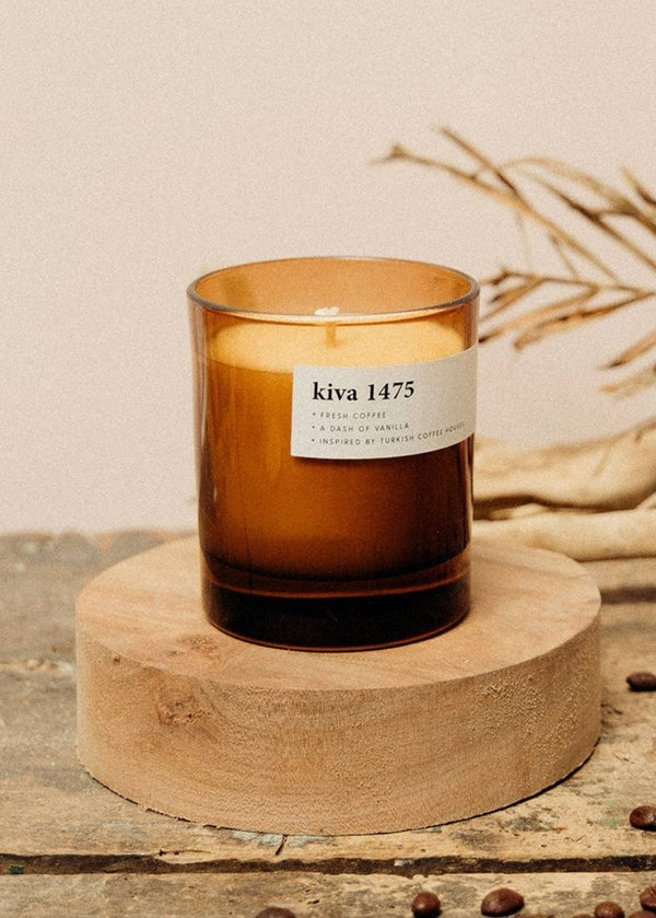 A brown soy wax candle with a label that reads 'KIVA 1475 THE COFFEE CANDLE' placed on a wooden candle holder, with coffee beans scattered around.