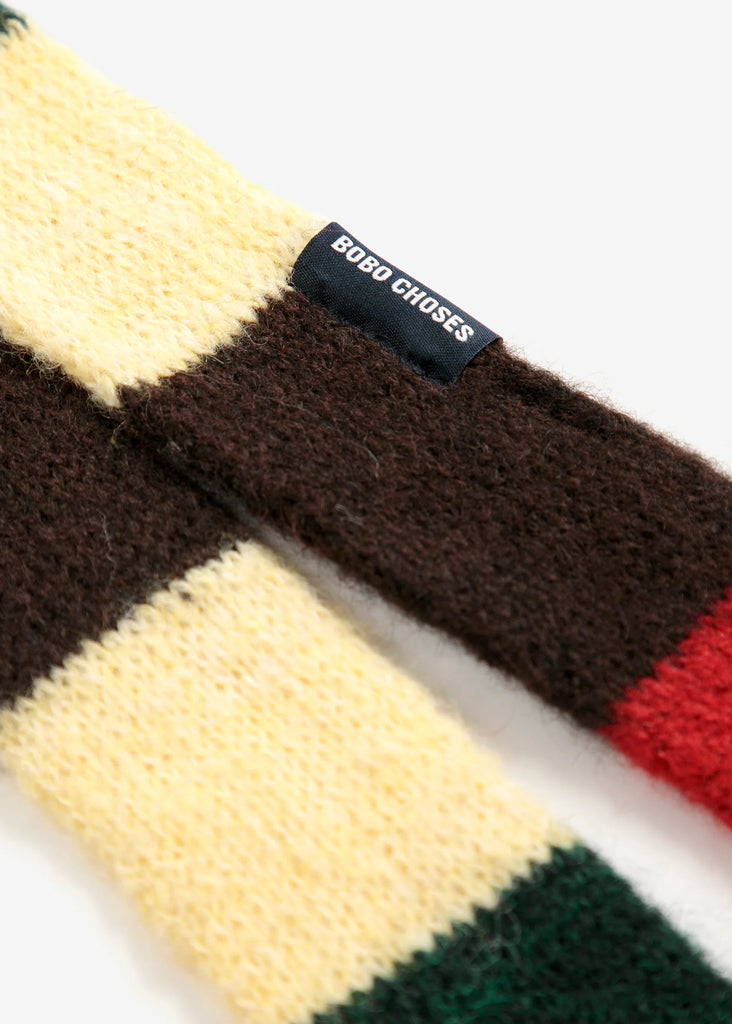 A close-up of a chunky knit striped accessory, likely a scarf or sweater, featuring blocks of cream, dark brown, red, and green, with a black fabric brand tag reading 'BORO CHOSES'.