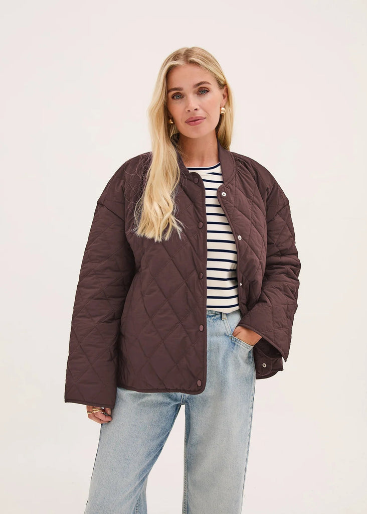 A blonde woman wearing a dark brown diamond-quilted puffer jacket over a black and white striped t-shirt and light-wash blue jeans, standing against a plain off-white studio background.