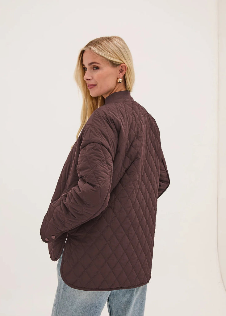 A woman with blonde hair is shown from the back wearing a dark brown diamond-quilted jacket and light blue jeans, standing against a plain white studio background.