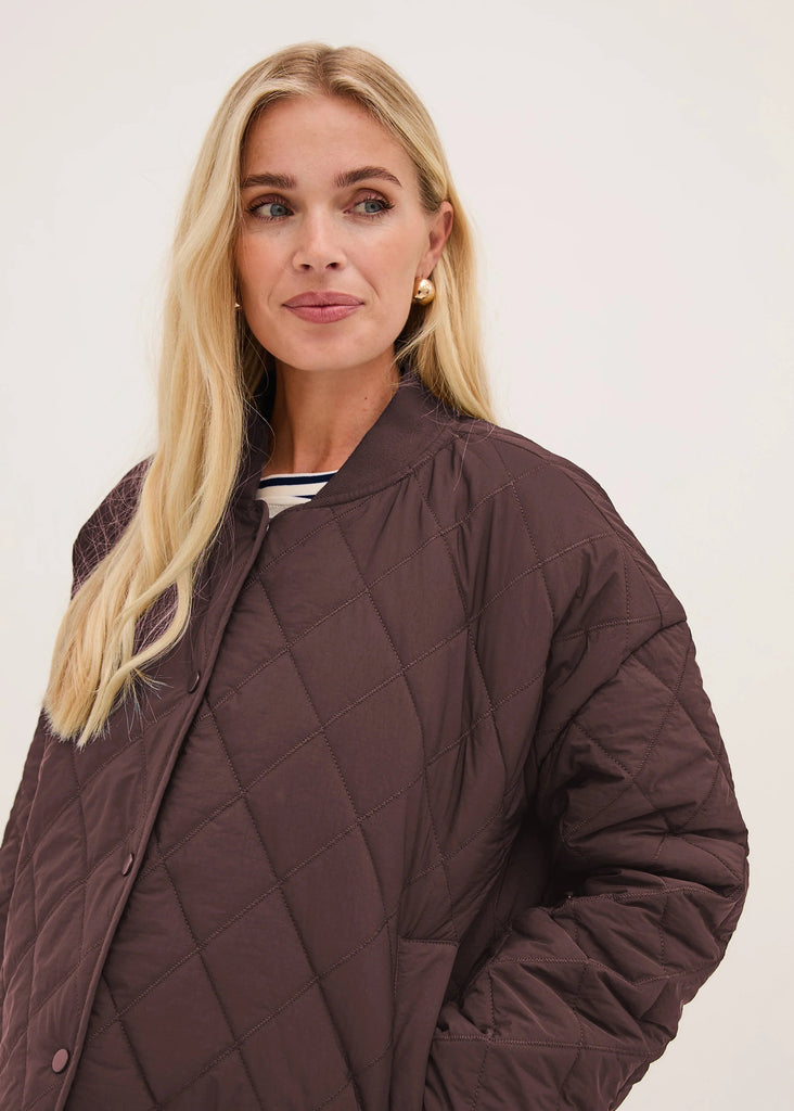 A blonde woman wearing a dark brown diamond-quilted jacket, photographed against a clean off-white studio background.