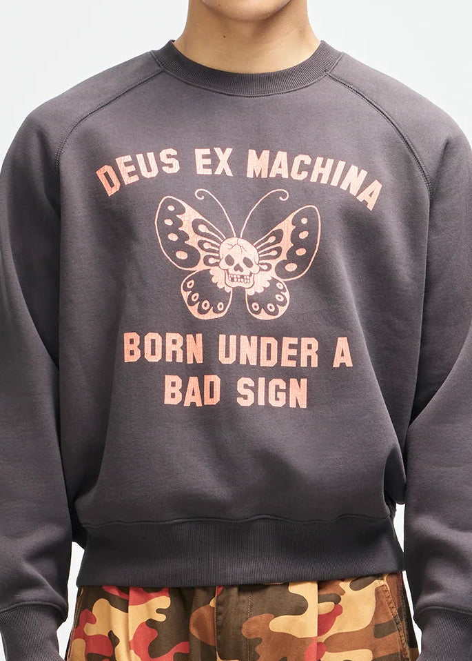 A model wearing a dark gray crewneck sweatshirt with a pink graphic of a butterfly and the text 'DEUS EX MACHINA' and 'BORN UNDER A BAD SIGN', paired with camouflage cargo pants, against a plain white background.