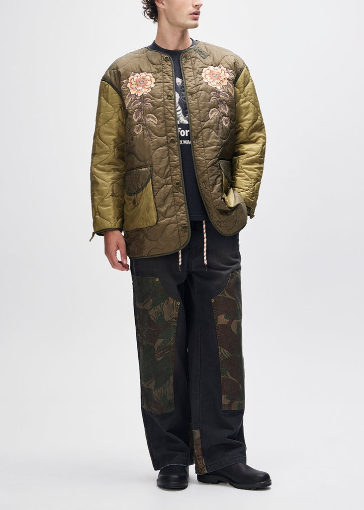 Person wearing a green quilted jacket with floral embroidery and camouflage pants on a white background