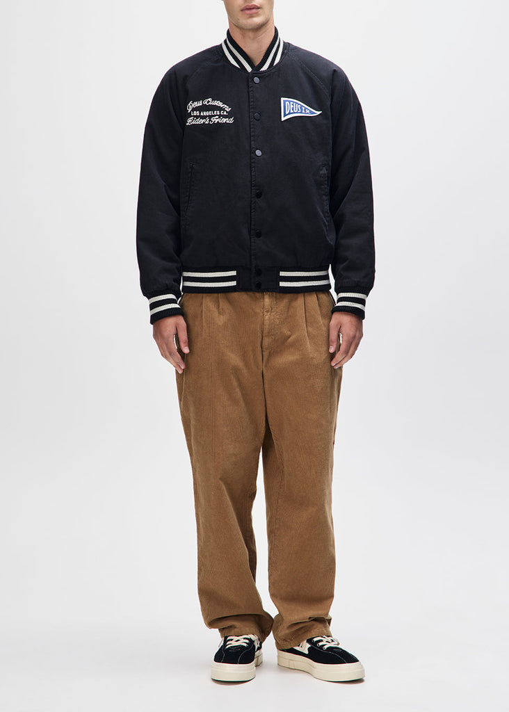 Person wearing a black varsity jacket with white accents and brown pants on a white background
